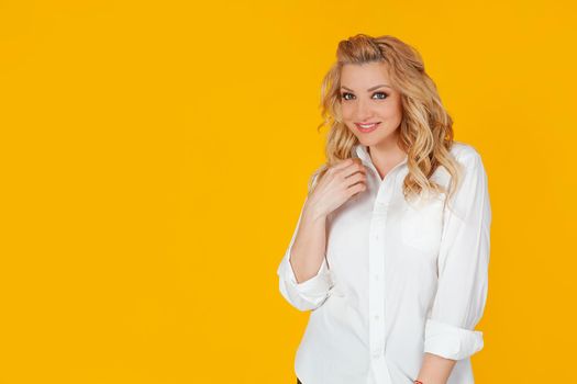 A blonde woman in a white shirt, neatly combing her hair with her hand, tilts her head and smiles affectionately at the camera with a coquettish, caring look, stands on a yellow background