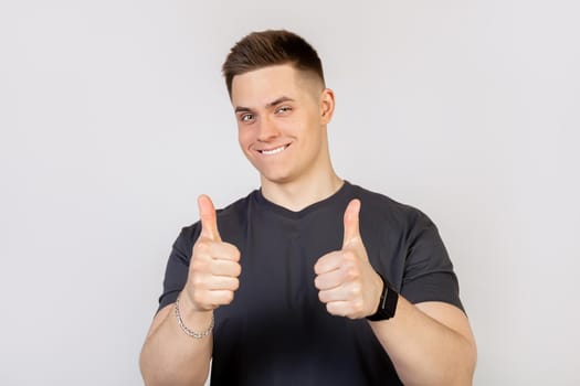A young man in a gray T-shirt on a white background, showing a finger up to the camera with a happy smile. All is well, shows success.