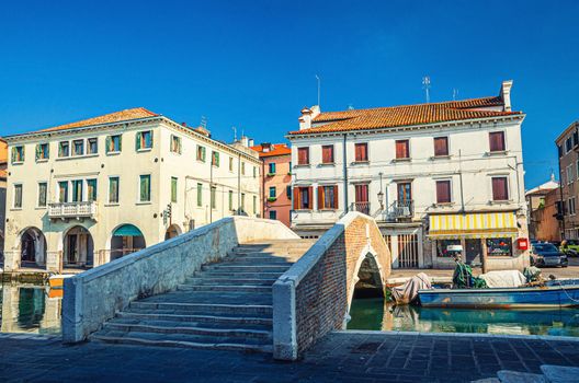 Stone brick bridge Ponte Pescheria across Vena water canal and old buildings in historical centre of Chioggia town, blue sky background in summer day, Veneto Region, Northern Italy