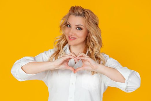 Blonde in white shirt, middle aged European girl showing heart gesture acknowledging sympathy, smiling friendly at camera, on yellow background.