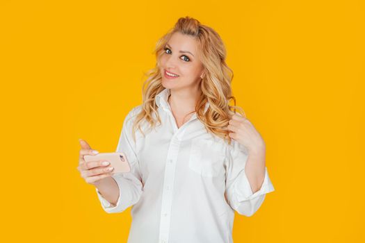 European middle-aged woman in a white shirt, pays attention to the camera, is distracted from the smartphone, smiles friendly and joyfully holds a mobile phone, standing on a yellow background