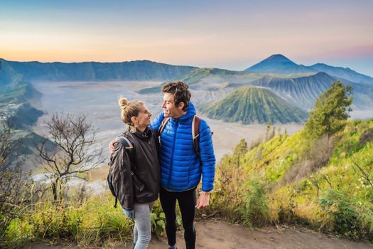Young couple man and woman meet the sunrise at the Bromo Tengger Semeru National Park on the Java Island, Indonesia. They enjoy magnificent view on the Bromo or Gunung Bromo on Indonesian, Semeru and other volcanoes located inside of the Sea of Sand within the Tengger Caldera. One of the most famous volcanic objects in the world. Travel to Indonesia concept.