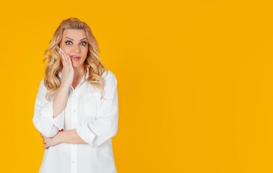 Woman blandin european middle-aged woman in white shirt, panic shocked upset, frowning, open mouth, touching her face with hand disappointed to hear bad news unhappy standing on yellow background.