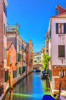 Venice cityscape with narrow water canal with boats moored between old colorful buildings and stone bridge, Veneto Region, Northern Italy. Typical Venetian view, vertical view, blue sky background