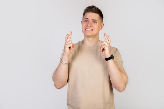 A handsome young guy crosses his fingers with a worried expression, asking God please, keep your fingers crossed with a pitiful look, hoping that the boss did not notice that he is late for work.
