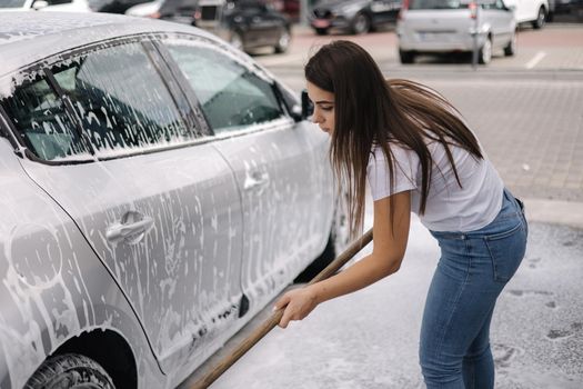 Attractive young woman washing her car with shampoo and brushes. Female washes automobile with foam and water outside on self service car wash.