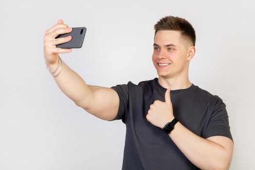 Portrait of a stylish handsome smiling young man taking a selfie on a smartphone, holding a mobile phone in his outstretched hand and trying to look beautiful in photo white background Online concept