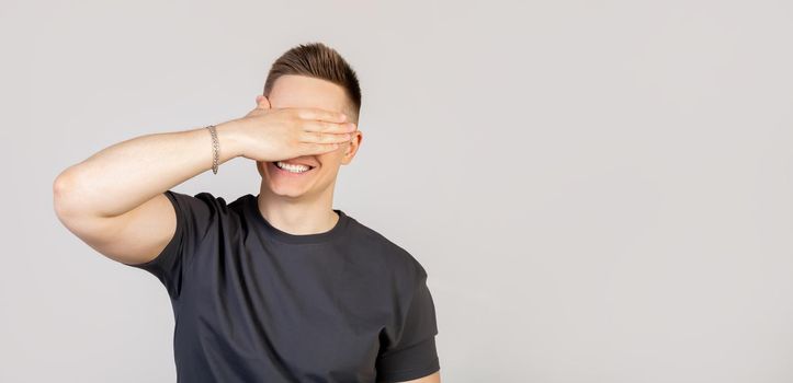 Portrait of a charming young man, an excited European guy, covering his eyes with his palm and smiling, waiting for an unexpected gift, waiting for a gift, counting ten, playing hide and seek