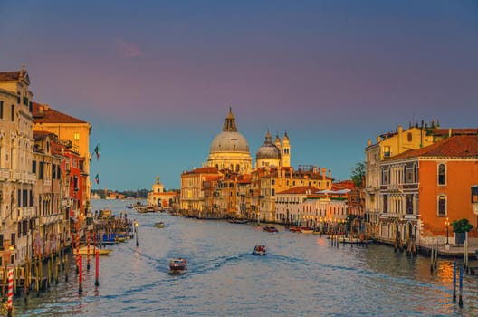 Venice cityscape with Grand Canal waterway. Buildings with evening lights along Grand Canal. Santa Maria della Salute Roman Catholic church on Punta della Dogana at twilight. Veneto Region, Italy.