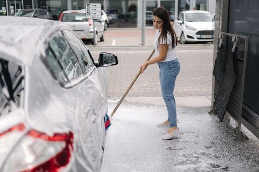 Attractive young woman washing her car with shampoo and brushes. Female washes automobile with foam and water outside on self service car wash.