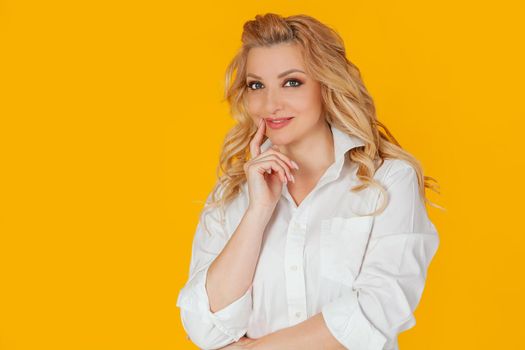 Attractive happy European blond woman in white shirt smiling curiously, touching her finger to her lips thoughtfully. Standing on a yellow background
