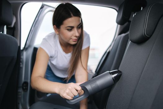 Attractive young woman using portable vacuum cleaner in her car. Woman vacuuming her car in the garage at home. Car interior cleaning.