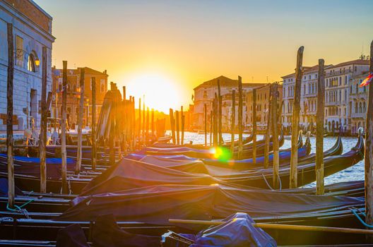 Gondolas moored docked on pier of Grand Canal waterway in Venice. Baroque style buildings along Canal Grande background. View against sun. Amazing Venice cityscape at sunset. Veneto Region, Italy