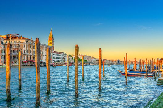 Venice evening view with wooden poles and boats near pier of Grand Canal waterway at sunset, Campanile bell tower and row of baroque style buildings in San Marco sestiere, Veneto region, Italy