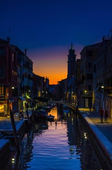 Venice cityscape with narrow water canal, fondamenta embankment. Silhouette of bridge, bell tower and buildings. Veneto Region, Northern Italy. Typical Venetian view at sunset evening, vertical view