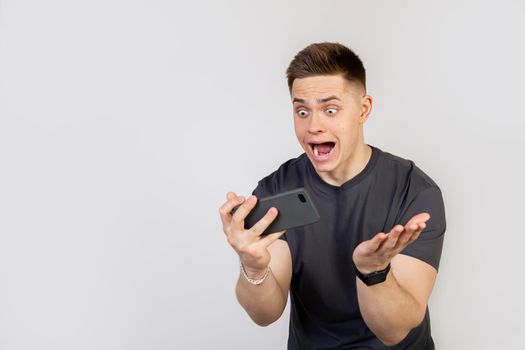 A surprised, shocked young man holds a smartphone in his hands and looks at the display with his mouth open and big eyes. A guy with a sagging jaw in fear came out first in the game, became a champion