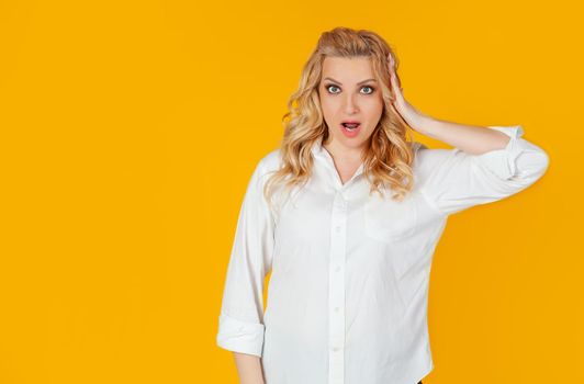 European young woman blonde in panic and shock upset, open mouth disappointed, touching her head upset to hear bad news, standing upset, yellow background