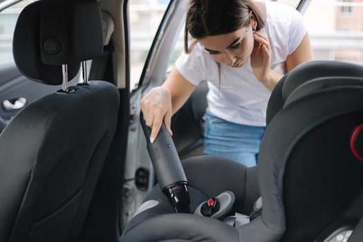 Close-up of female using portable vacuum cleaner in her car. Baby ca seat cleaning. Woman vacuuming seats. Dust and dirt removal.