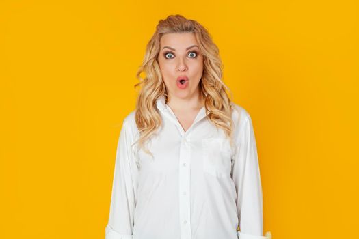 An excited impressed woman, a blonde in a white shirt, with a surprised open mouth, looks frightened at the camera, expressing amazement from the shock. Isolated on a yellow background.