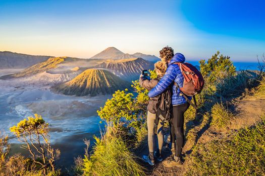 Young couple man and woman meet the sunrise at the Bromo Tengger Semeru National Park on the Java Island, Indonesia. They enjoy magnificent view on the Bromo or Gunung Bromo on Indonesian, Semeru and other volcanoes located inside of the Sea of Sand within the Tengger Caldera. One of the most famous volcanic objects in the world. Travel to Indonesia concept.