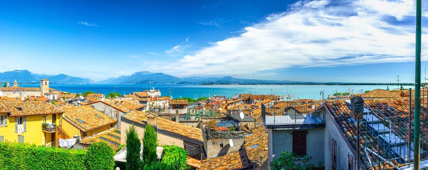 Panorama of Desenzano del Garda town with red tiled roof buildings, Garda Lake water, Monte Baldo mountain range, Sirmione peninsula, Lombardy, Northern Italy. Aerial panoramic view of Desenzano