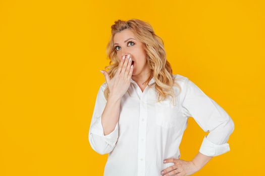 Portrait of a woman in a white shirt, a middle-aged European girl, yawning and covering her open mouth, with a boring face, wakes up early, stands on a yellow background.