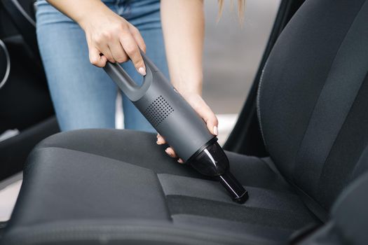 Close up of female using portable vacuum cleaner in her car. Electrical vacuum in woman's hand clean car inside from dust.