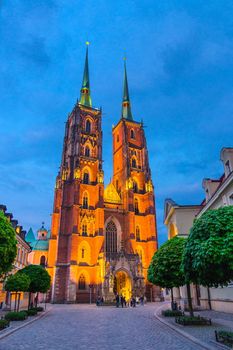 Cathedral of St. John the Baptist catholic church building with two spires and lights, cobblestone road street with green trees in old town historical city centre of Wroclaw, evening view, Poland