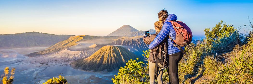 BANNER, LONG FORMAT Young couple man and woman meet the sunrise at the Bromo Tengger Semeru National Park on the Java Island, Indonesia. They enjoy magnificent view on the Bromo or Gunung Bromo on Indonesian, Semeru and other volcanoes located inside of the Sea of Sand within the Tengger Caldera. One of the most famous volcanic objects in the world. Travel to Indonesia concept.