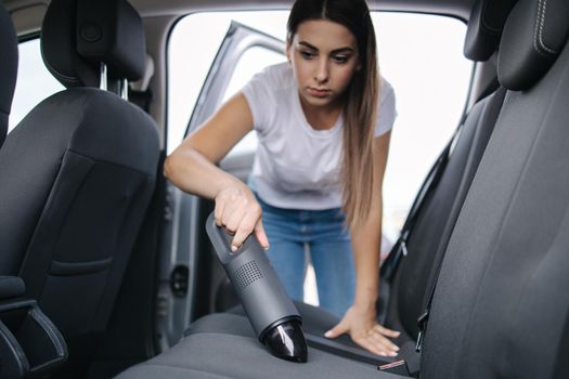 Attractive young woman using portable vacuum cleaner in her car. Woman vacuuming her car in the garage at home. Car interior cleaning.