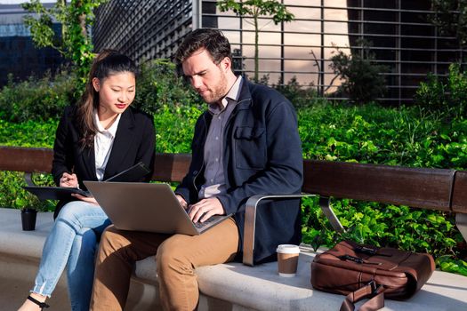 asiatic woman and caucasian man working together with laptop in a park next to the office, concept of coworkers and diversity at work, copy space for text