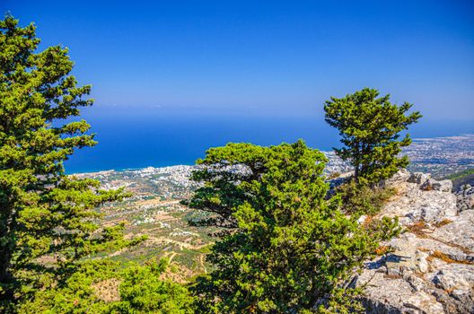 Aerial view of Kyrenia District valley with Kyrenia Girne city and Mediterranean sea in sunny day, green trees on rock of Kyrenia Girne mountains foreground, blue sky background, Northern Cyprus