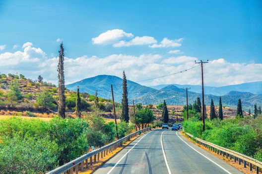 Landscape of Cyprus with cars vehicles riding asphalt road in valley with yellow dry fields, cypress trees and roadside poles, Troodos mountain range and hills, clear blue sky in sunny day background