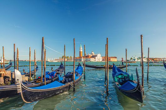 Gondolas moored docked on water in Venice. Gondoliers sailing San Marco basin waterway. San Giorgio Maggiore island with Campanile San Giorgio in Venetian Lagoon, blue clear sky, Veneto Region, Italy