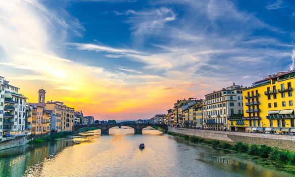 St Trinity Bridge stone bridge and boat on Arno River water and embankment promenade with buildings in historical centre of Florence city, bright blue orange evening sky clouds, Tuscany, Italy