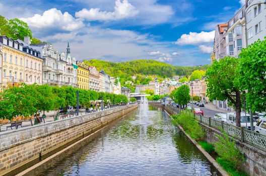 Karlovy Vary Carlsbad historical city centre with Tepla river central embankment, colorful beautiful buildings, Slavkov Forest hills, blue sky white clouds background, West Bohemia, Czech Republic