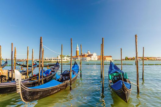 Gondolas moored docked on water in Venice. Gondoliers sailing San Marco basin waterway. San Giorgio Maggiore island with Campanile San Giorgio in Venetian Lagoon, blue clear sky, Veneto Region, Italy