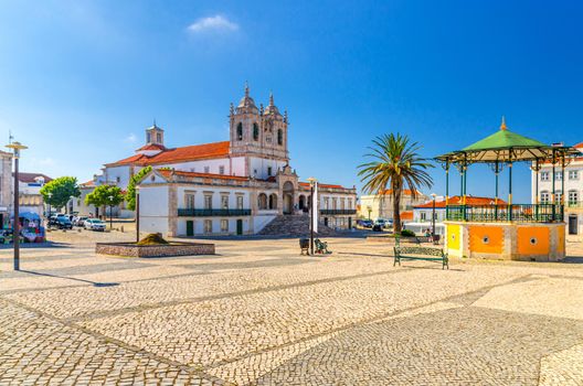 Sanctuary of Our Lady of Nazare catholic church in cobblestone square with palm trees in Sitio hilltop da Nazare town, clear blue sky in sunny summer day, Leiria District, Oeste region, Portugal