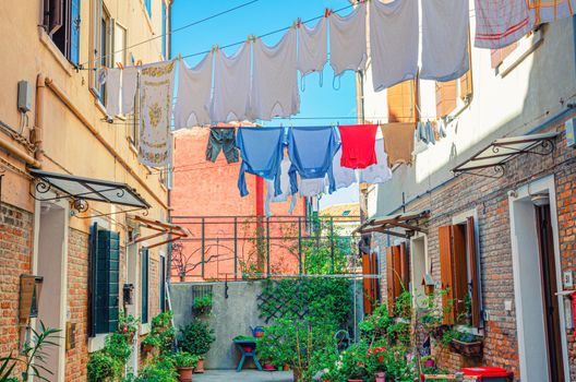 Typical italian courtyard between buildings with brick walls, green plants and flowers in flowerpot, small tables, wet clothes hanging on cord, Murano islands, Veneto Region, Northern Italy