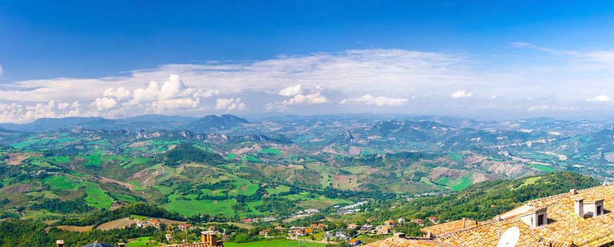 Aerial top panoramic view of landscape with valley, green hills, fields and villages of Republic San Marino suburban district with blue sky white clouds background. View from San Marino fortress.