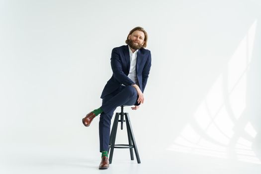 a handsome brutal bearded and long-haired man, a stylish businessman sitting on a chair with a white background. High quality photo