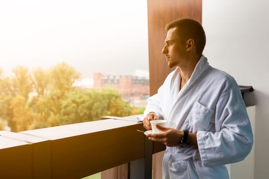 A young man in a white coat on the balcony with a cup of coffee. Morning coffee or tea is holding a cup of man on the summer terrace