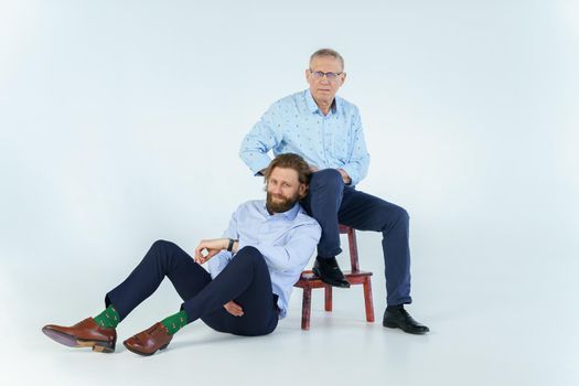 Father sits on the chair and son sits on white background, both men look into the camera, an elderly man in glasses with diopters, a stylish young man with a beard and long hair. High quality photo