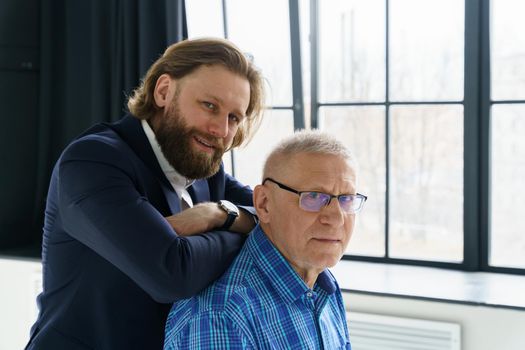 the son in a stylish suit leans on the shoulders of his father, Father and son in huge window background, both men look into the camera, an elderly man in glasses, young man with a beard and long hair. High quality photo