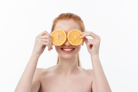 Portrait of a cheerful young girl holding two slices of an orange at her face over white wall background.