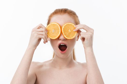Portrait of a cheerful young girl holding two slices of an orange at her face over white wall background.