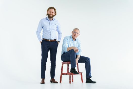 Father sits on the chair and son stands on white background, both men look into the camera, an elderly man in glasses with diopters, a stylish young man with a beard and long hair. High quality photo