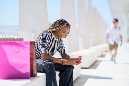 Young black woman using her smartphone sitting on a bench in the street.