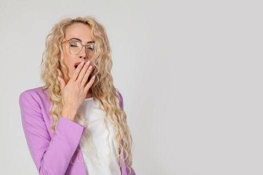 Snapshot of a sleepy attractive Caucasian woman blonde with curly hair wearing glasses, feeling tired after a night without sleep, yawning, covering her open mouth with her palm