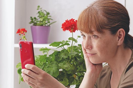 Middle aged caucasian woman making selfie on phone or chatting indoor on Geranium flowers background